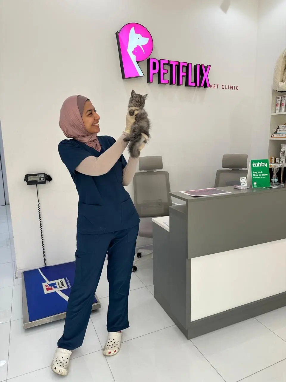 Female vet in navy scrubs and hijab lifting a kitten in reception under PETFLIX neon sign, scale and desk visible
